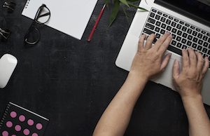 Workspace with laptop, male hands, notebook,eyeglasses, sketchbook, black wooden desk with bamboo leaf . Flat lay, top view office table desk. Freelancer working place.